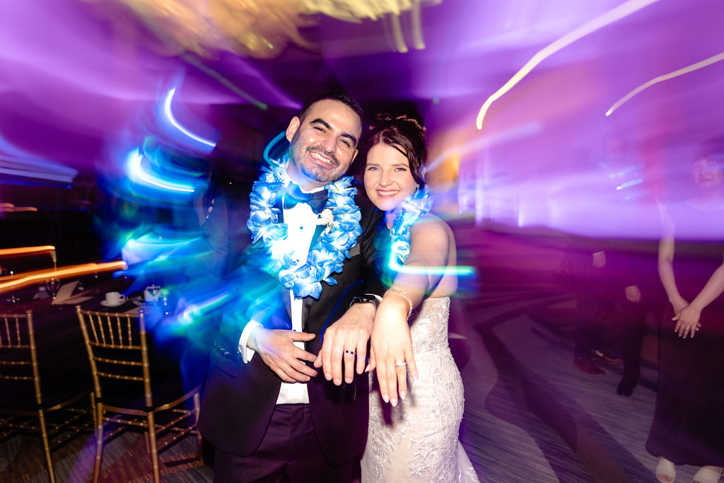 Bride and groom celebrating with blue leis under colorful reception lights