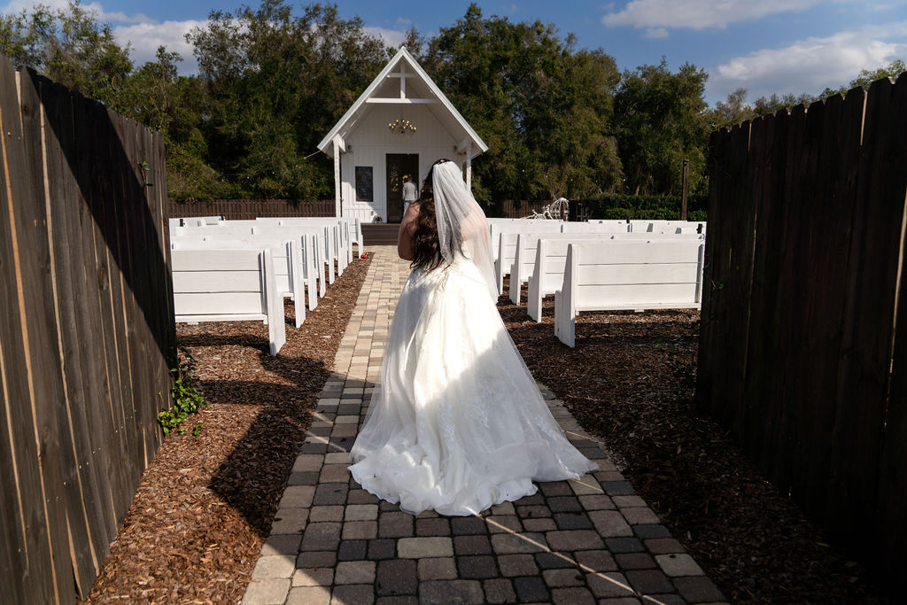 Bride walking alone along an outdoor aisle toward a white chapel