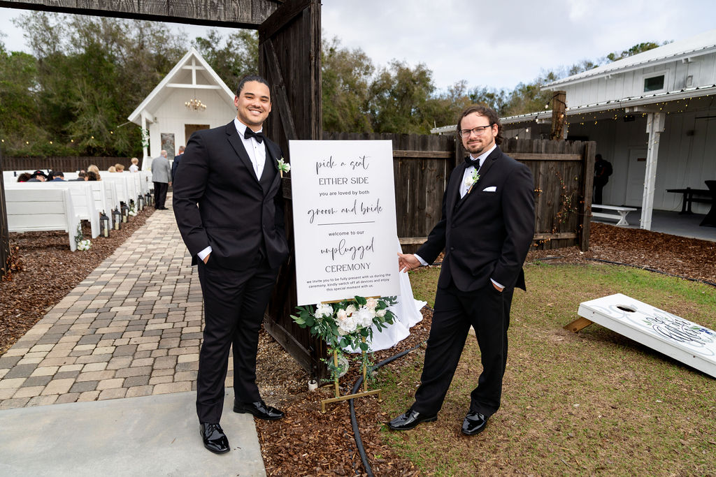 Two men in tuxedos beside an outdoor wedding welcome sign