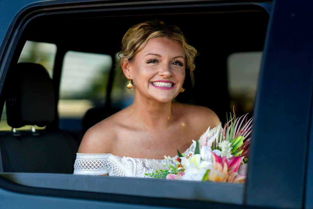 Bride smiling from the window of a pickup truck before a waterfront wedding