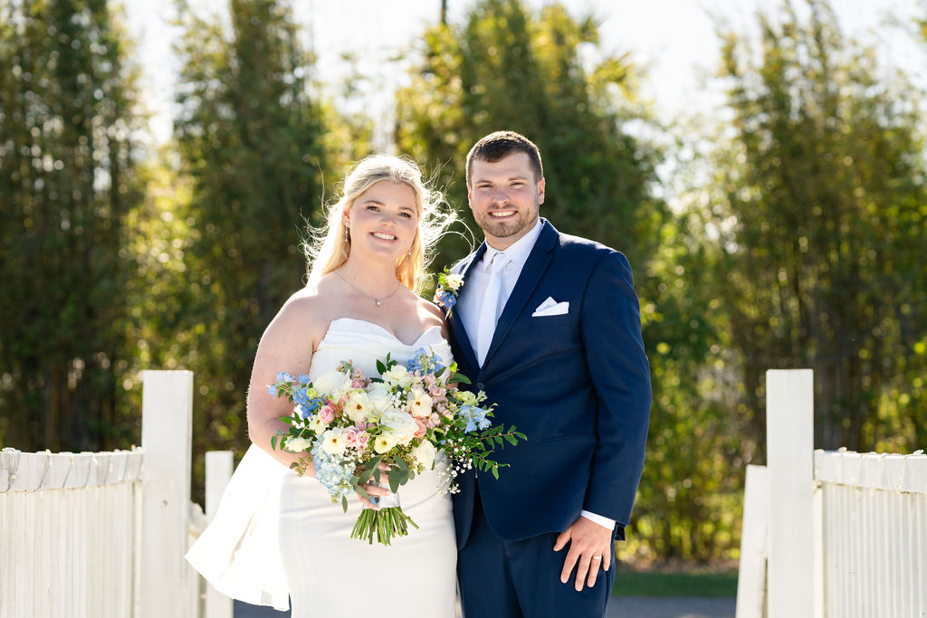 Bride and groom posing with a colorful bouquet beside a white fence