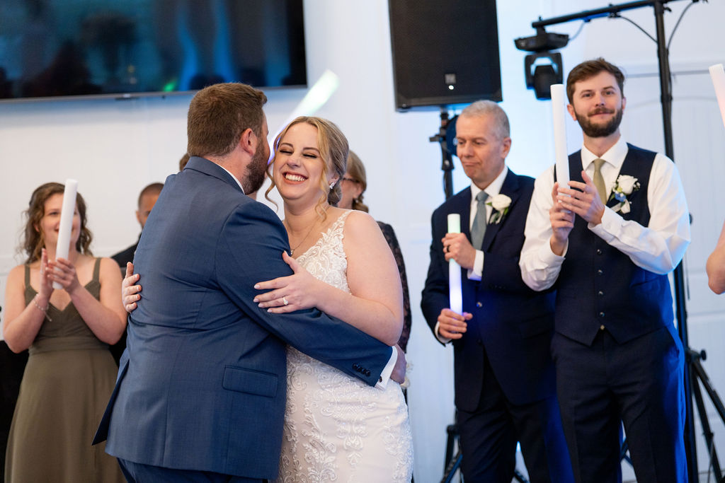Bride and groom embracing as adult guests applaud, with a recording camera visible behind them