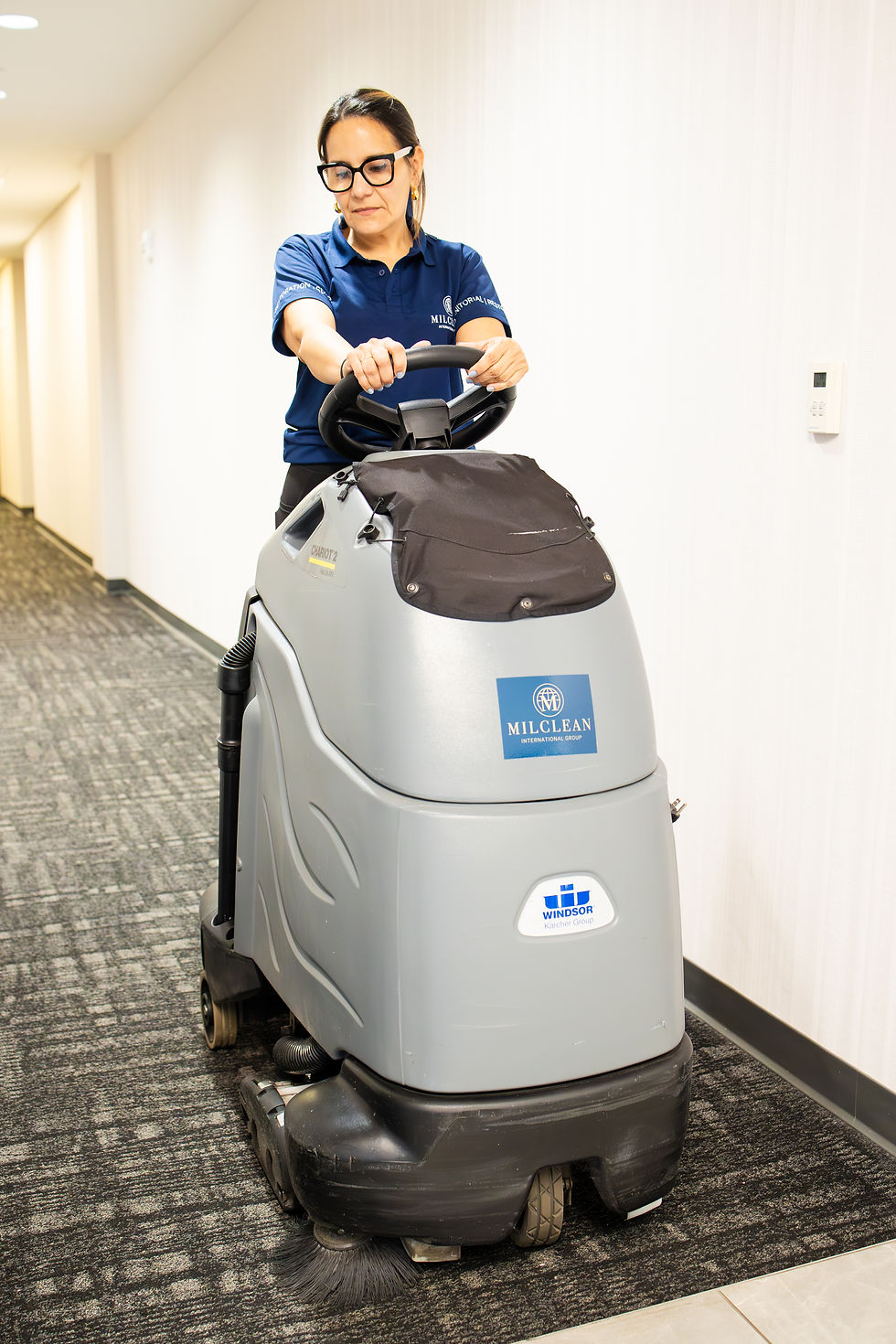 A uniformed team member operates a MilClean floor-care machine along a carpeted corridor.
