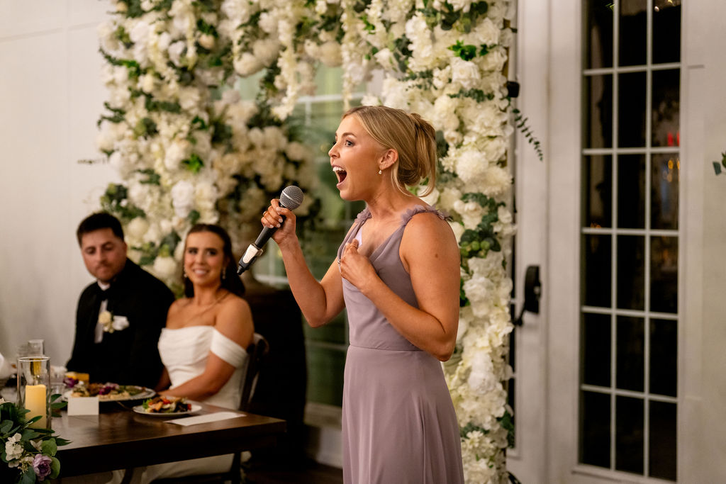 Bridesmaid giving a toast beside a floral reception backdrop