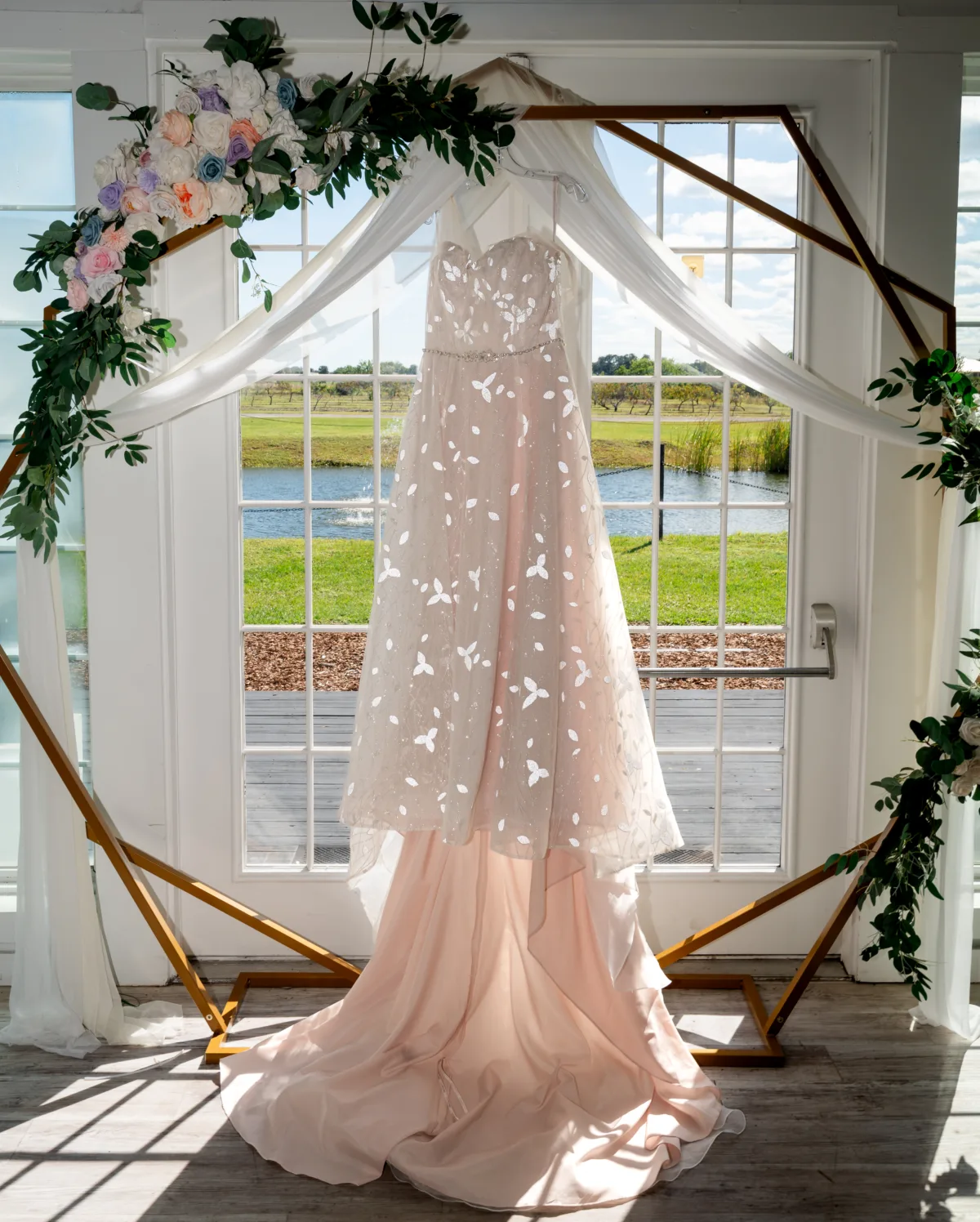 Elegant wedding dress hanging in front of window with floral ceremony arch accents