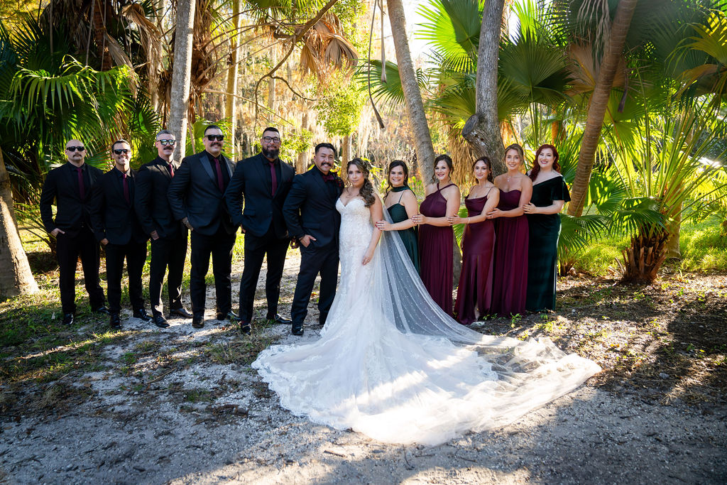 Wedding party posing together beneath palms with the bride’s dress train spread out