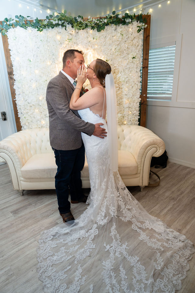 Full-length bride and groom kiss with a lace train beside a cream sofa