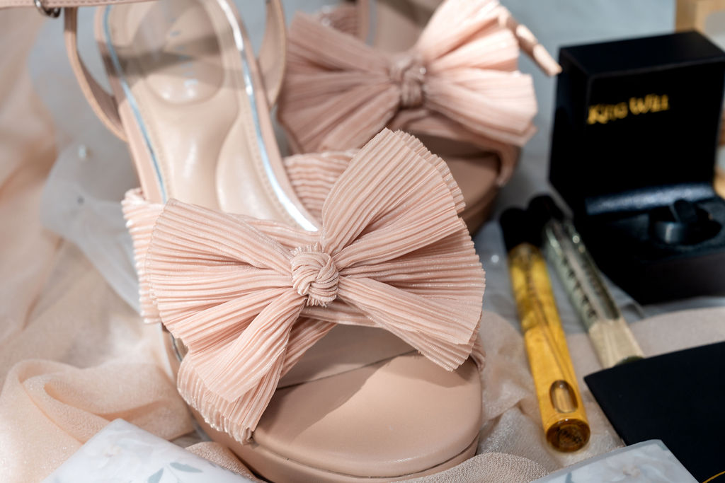 Blush pink bow-front wedding shoes beside perfume and a ring box