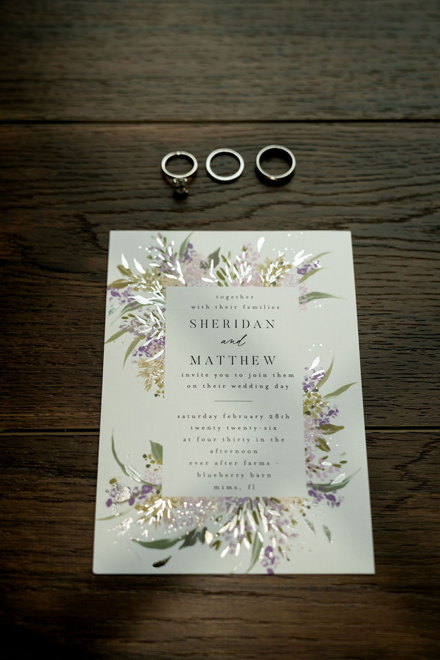 Three rings above a floral wedding invitation on a wooden surface