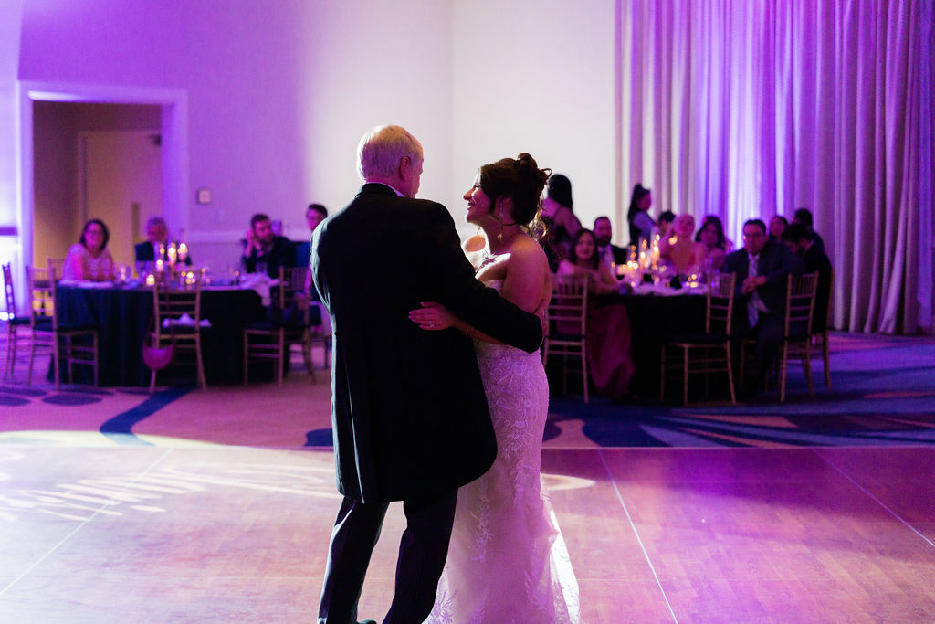 Bride dancing with her father beneath purple reception lights
