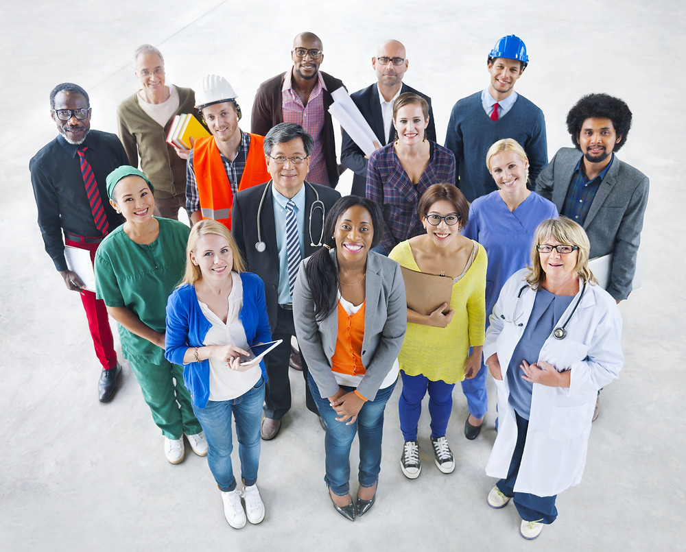 A group of fifteen adult men and women of diverse ages and ethnicities, wearing clothing and holding props associated with diverse professions (technology, medicine, construction, architecture, education, etc.), stand smiling and looking up at the camera.