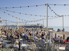 Outdoor restaurant with people, pier in the background, sunny day, clear blue sky.
