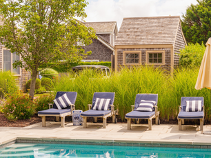 Four blue lounge chairs with striped pillows face a pool, set against lush green grass and rustic wooden cottages under a sunny sky.