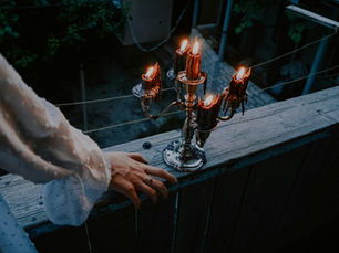 A hand rests on a rustic wooden railing beside a lit candelabra with six flickering candles. Dark, lush foliage forms the background.