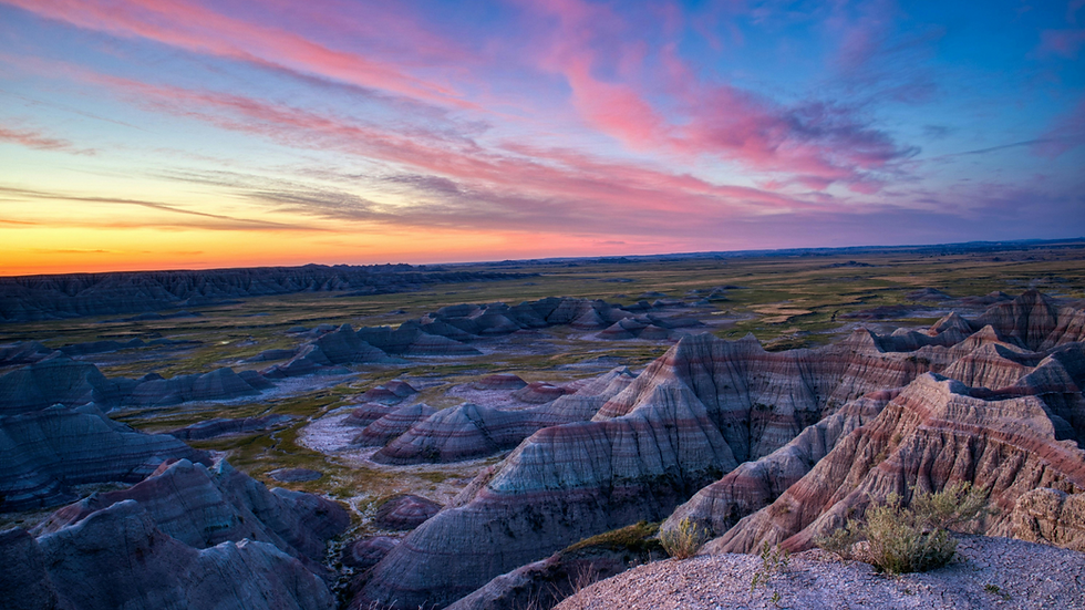 Colorful sunset over Badlands, featuring layered rock formations and vast landscape. Sky painted with pink, orange, and blue hues.