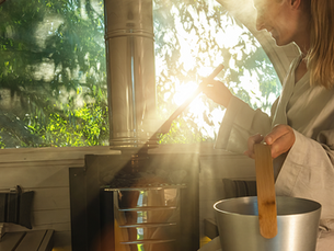 Person in a sauna, wearing a robe, stirs a stove with a wooden ladle. Sunlight filters through a large window, showing lush greenery.