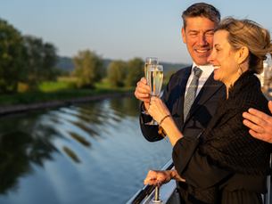 A couple in formal attire toasts with champagne on a boat, smiling and enjoying a sunny river view with greenery in the background.