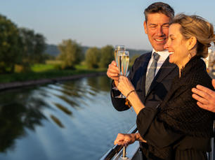 A couple in formal attire toasts with champagne on a boat, smiling and enjoying a sunny river view with greenery in the background.