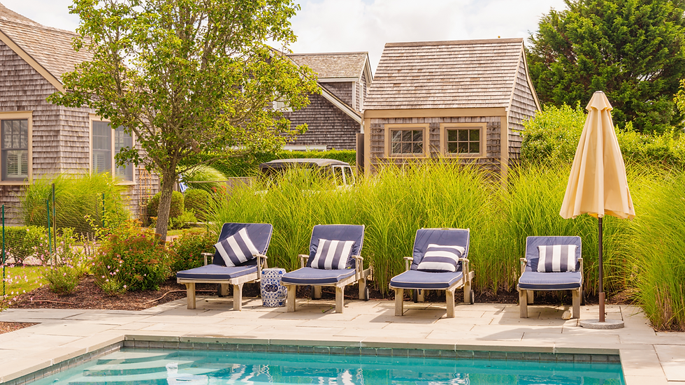 Four blue lounge chairs with striped pillows face a pool, set against lush green grass and rustic wooden cottages under a sunny sky.