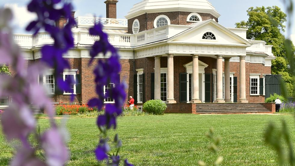 Historic brick building with columns, seen through vibrant purple flowers. Two people walk in the garden. Bright, sunny day.