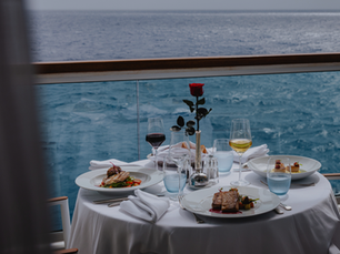 Elegant dining setup on a balcony overlooking the ocean. Two plates of gourmet food, wine glasses, and a rose centerpiece create a romantic mood.