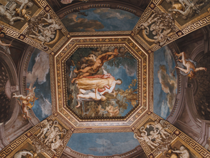 Ornate palace ceiling fresco with classical mythological figures in gold frames and blue sky panels, viewed from below.