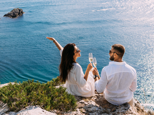 Couple in white sit on a rocky cliff, toasting with champagne above a sparkling blue sea on a sunny day.