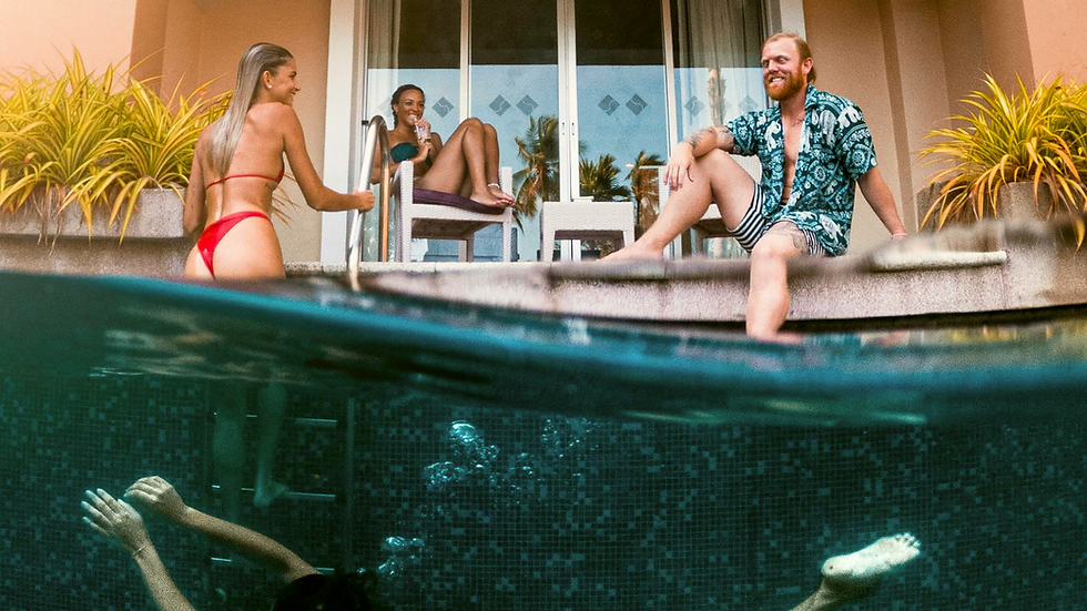 Three people relax by a pool. A woman in a red bikini climbs out, another lounges on a chair, and a man smiles. Below, someone swims underwater.