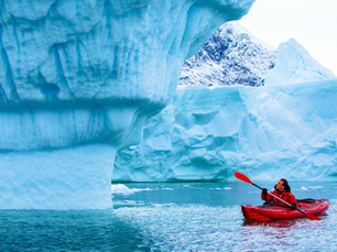 Person kayaking in a red kayak near large blue icebergs, looking up, surrounded by icy water, with a snowy mountain in the background.