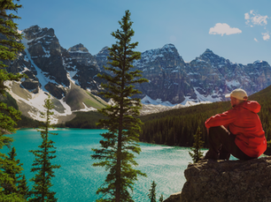 Person in red jacket and cap sits on rock overlooking turquoise lake. Snowy mountains and pine trees surround, under clear blue sky.