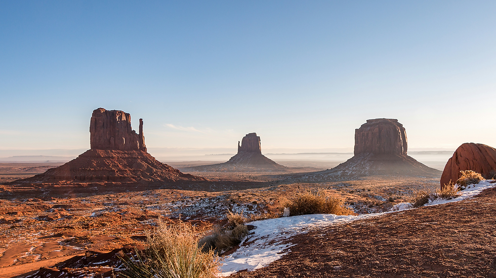 Desert landscape with three towering rock formations under a clear blue sky. Snow patches and shrubs are visible on the reddish-brown ground.