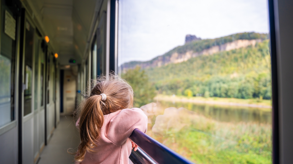 Child in pink hoodie gazes out train window at green landscape and cliffs. The interior is cozy, creating a peaceful and curious mood.