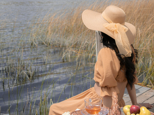 Woman in a wide-brimmed hat sits on a wooden dock by a lake, with fruit and tea beside her. Tall grass surrounds the serene setting.