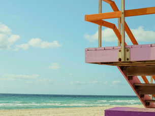 Colorful lifeguard tower with pink and orange railings by a sandy beach. Blue sky and ocean waves create a serene, sunny atmosphere.