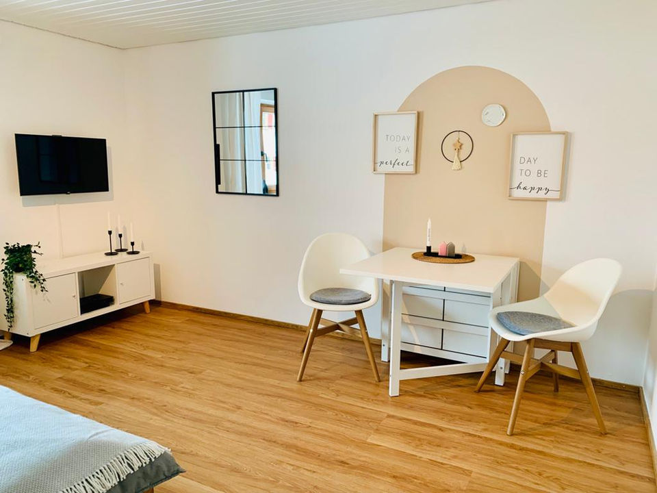 Ausstattung Appartement Kleine Auszeit: Gemütliche Essecke mit vergrößerbarem Tisch und zwei Stühlen. Auf der anderen Seite, Flachbildfernseher mit TV-Board. In diesem befindet sich eine Auswahl an Büchern, Wanderkarte und Fahrradkarte passend zum Allgäu.