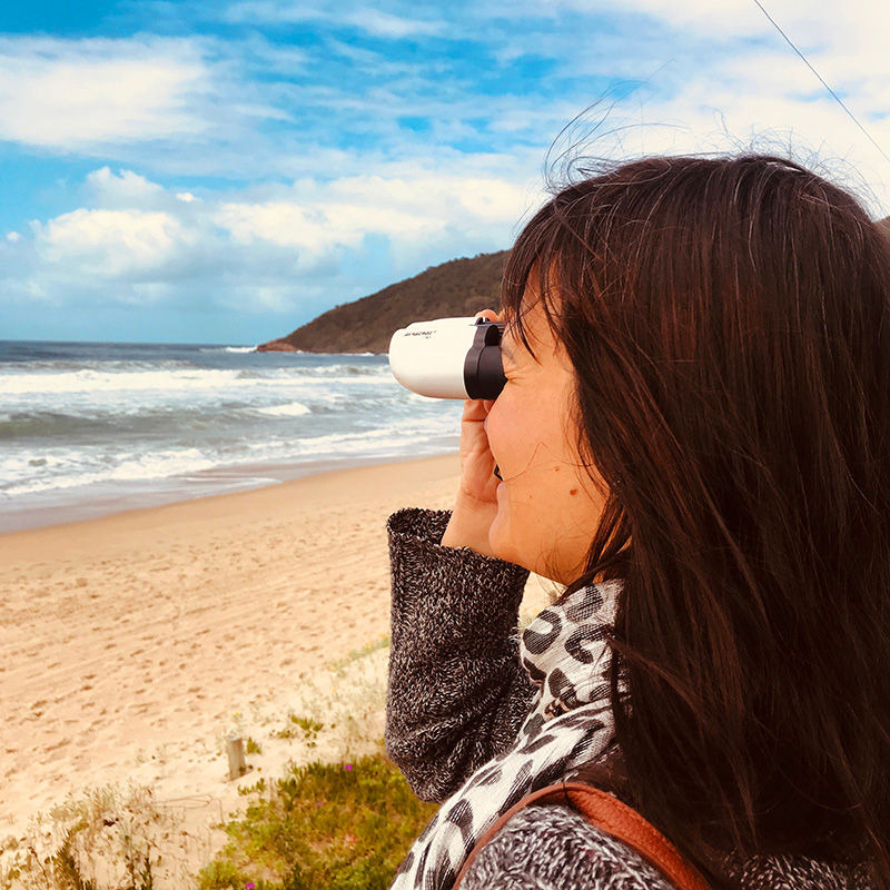 Mulher com binóculos avistando a baleia franca na beira da praia.