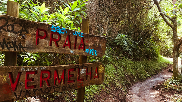 Placa escrito Praia Vermelha e com a trilha e mata ao fundo.