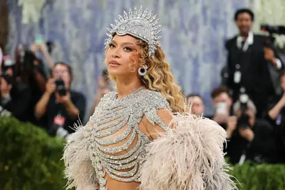 Beyoncé at the Met Gala 2026 wearing a crystal-embellished, sculptural gown with a dramatic feathered coat and ornate headpiece, posing confidently on the red carpet against a backdrop of photographers.