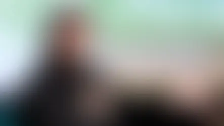 Kenneth Cole speaking on stage at a conference, holding a microphone and gesturing during a panel discussion, with a blurred presentation screen in the background.