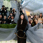 Madonna arrives at the 2026 Met Gala in a dramatic black look with a flowing sheer cape and a striking ship-inspired headpiece, surrounded by photographers and guests on the red carpet.
