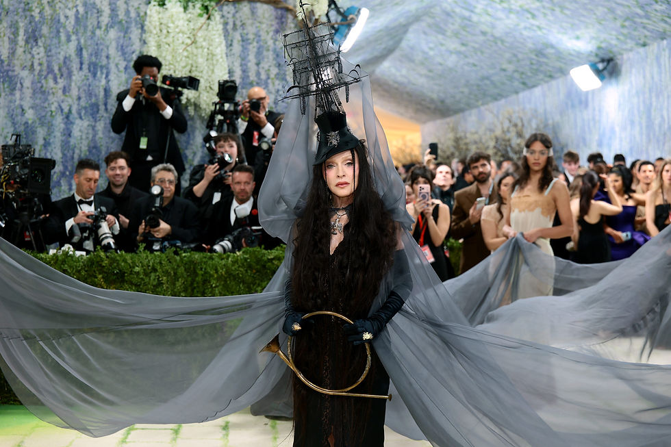Madonna arrives at the 2026 Met Gala in a dramatic black look with a flowing sheer cape and a striking ship-inspired headpiece, surrounded by photographers and guests on the red carpet.