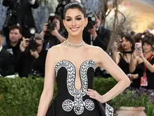 Anne Hathaway smiles on the 2026 Met Gala red carpet wearing an elegant black strapless gown with artistic white detailing, paired with sparkling diamond jewelry and a sleek bun hairstyle.
