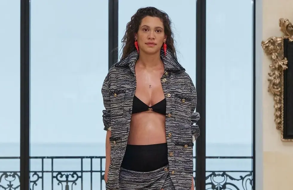 Pregnant model Kaya Wilkins walks the Chanel Resort 2027 runway wearing a black and white tweed skirt suit styled with a black bikini top, carrying a chain handbag, set against a bright seaside backdrop with large windows.