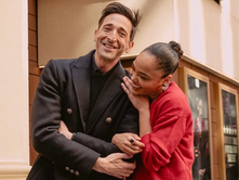 Two people smiling and walking together on a city street, sharing a warm, candid moment in stylish outfits.