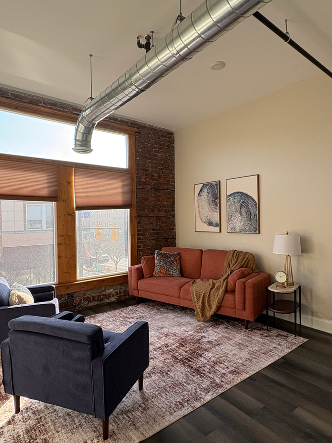 Office view of Downtown Port Huron in our counseling office. Reclaimed hardwood floors, large ornamental rectangle rug, two large windows providing a view of Port Huron's main street. Two comfortable chairs arranged across from one large couch which perpendicular to the large windows. Decorative pillows included on couch with small side table and lamp. Exposed hvac and high ceilings. Natural scenic art in muted colors on the walls above the couch.