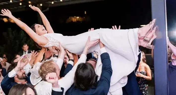 Bride crowd-surfed by guests at outdoor wedding reception with string lights, experiential event entertainment