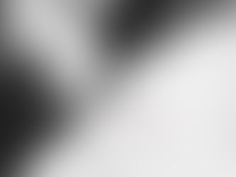 Black-and-white portrait of Goddess Becky reclining on a bed, wearing a sleek latex outfit. Her arms are raised above her head, framing her face, with long hair spread softly across the pillow. She gazes directly at the camera with a calm, self-assured expression, conveying quiet confidence and introspective energy.