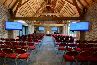 An empty conference room with red chairs arranged in theater-style seating facing a stage with a podium. Three large screens display "Genootschap van Notarissen West-Vlaanderen Van harte welkom." The room features a high wooden ceiling, brick walls, and is well-lit with natural light.
Tags:
Conference Room
Genootschap van Notarissen West-Vlaanderen
Formal Event
Professional Setup
Aventix Services
Theater-Style Seating
High Wooden Ceiling
Brick Walls
Event Preparation
Natural Lighting