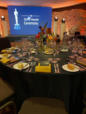 A table setting at The PLATO Awards Ceremony with wine glasses, plates with bread rolls, cutlery, yellow napkins, floral centerpieces, and small lamps. The background features a stage with a screen displaying the event's name and a trophy statue, creating a formal and prestigious atmosphere.
Tags:
PLATO Awards Ceremony
Elegant Table Setting
Wine Glasses
Floral Centerpieces
Sophisticated Dining
Prestigious Event
Brick Wall Backdrop
Award Trophy
Yellow Napkins
Event Lighting