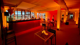 Cozy bar lounge with red-lit bar counter, bottles, glasses, and candles on tables. Two people present, one behind the bar, the other walking. Large windows and ambient lighting create a warm, inviting atmosphere, perfect for events or gatherings.
Tags:
Bar Lounge
Ambient Lighting
Cozy Atmosphere
Event Space
Red Lights
Elegant Bar
Intimate Setting
Large Windows
Indoor Venue
Lively Environment